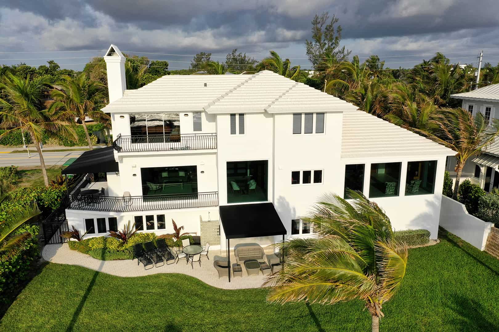 Drone view of large white home with black trim, three stories, front view, residential neighborhood, multiple double panel impact windows, FS-300 Maxi View Impact Storefront windows by Aldora, manicured lawn, palm trees, tropical environment