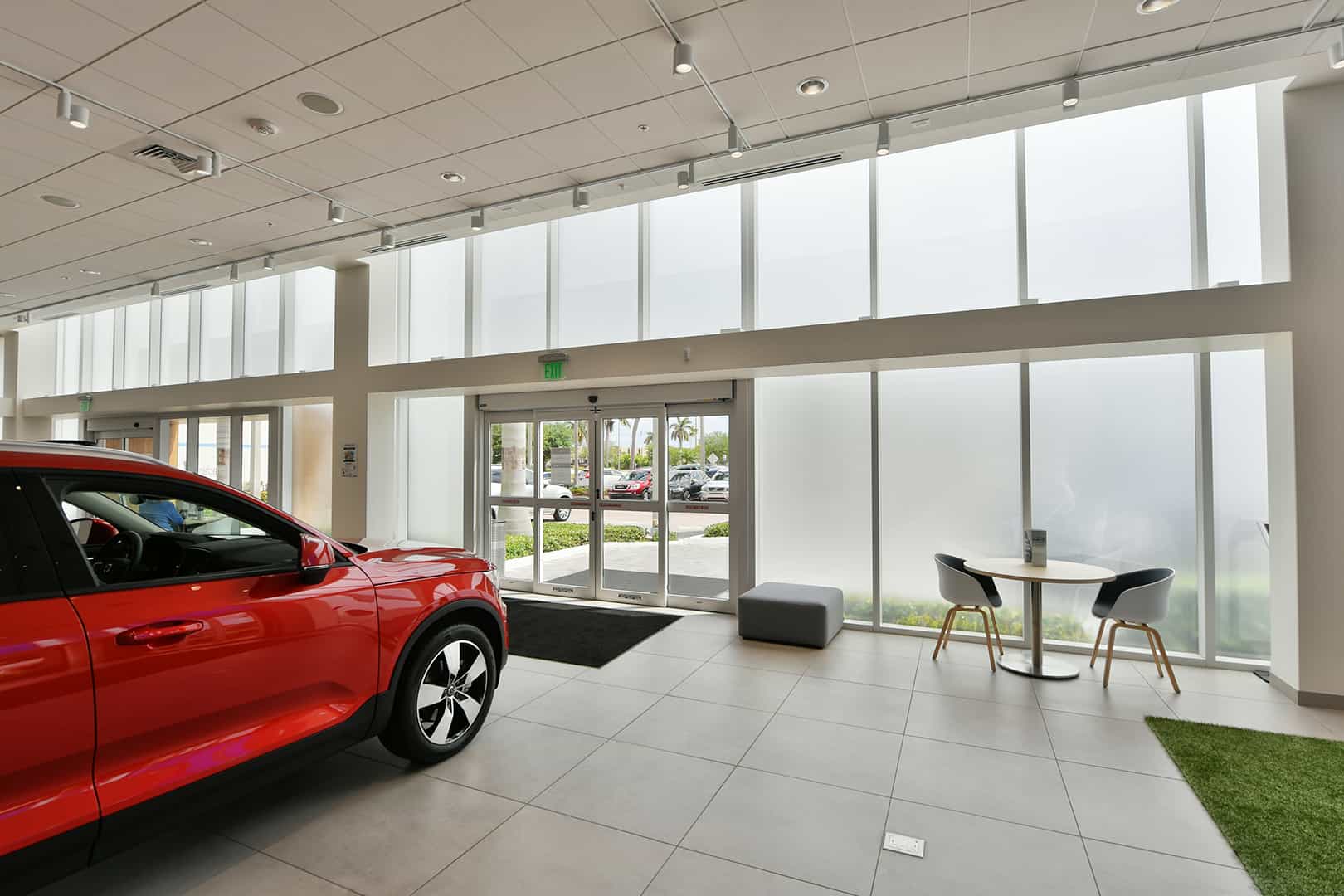 Interior view of car dealership looking through frosted FS-300 Maximum View Impact Storefront windows by Aldora, two sets of sliding glass doors, white tile floor and ceiling, seating area, red showcase vehicle in room