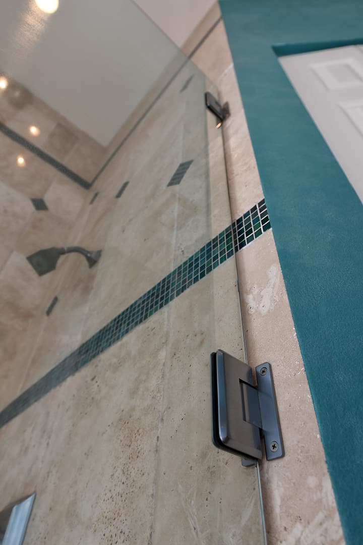 Angled close up view looking up at glass shower wall by Aldora, black burnished wall hinges, beige stone ceramic tile with green tile accents