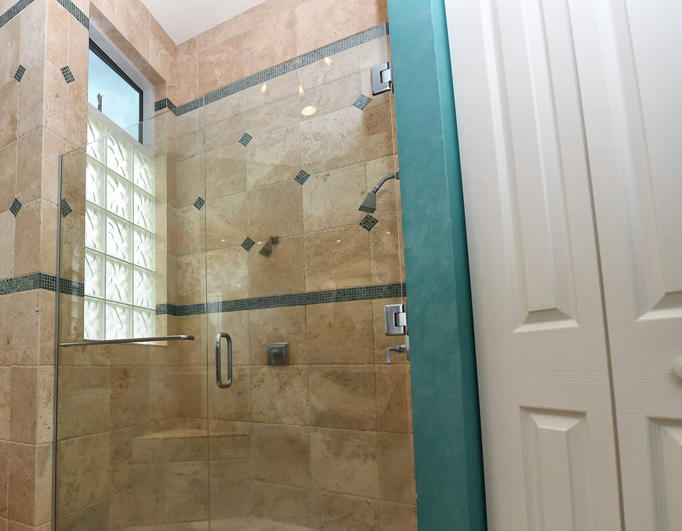 Floor view looking upward at master bathroom, beige ceramic tile with green accents, step up soaker tub, tiled glass window, accent candles, swinging glass door and glass shower wall by Aldora, chrome hardware