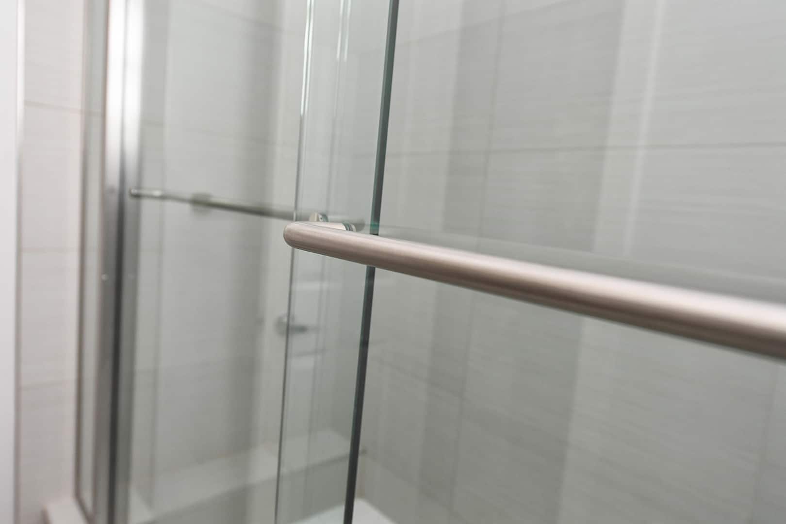 Angled close up view of chrome shower bar attached to sliding glass shower door by Aldora, slate grey tile in background