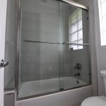 Looking up at sliding shower door by Aldora, chrome bar hardware attached the glass panels, chrome trim around glass shower walls, grey ceramic tile on floor and shower wall, standard white tub, toilet to right of shower