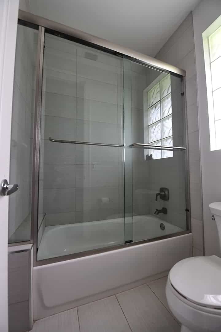 Looking up at sliding shower door by Aldora, chrome bar hardware attached the glass panels, chrome trim around glass shower walls, grey ceramic tile on floor and shower wall, standard white tub, toilet to right of shower
