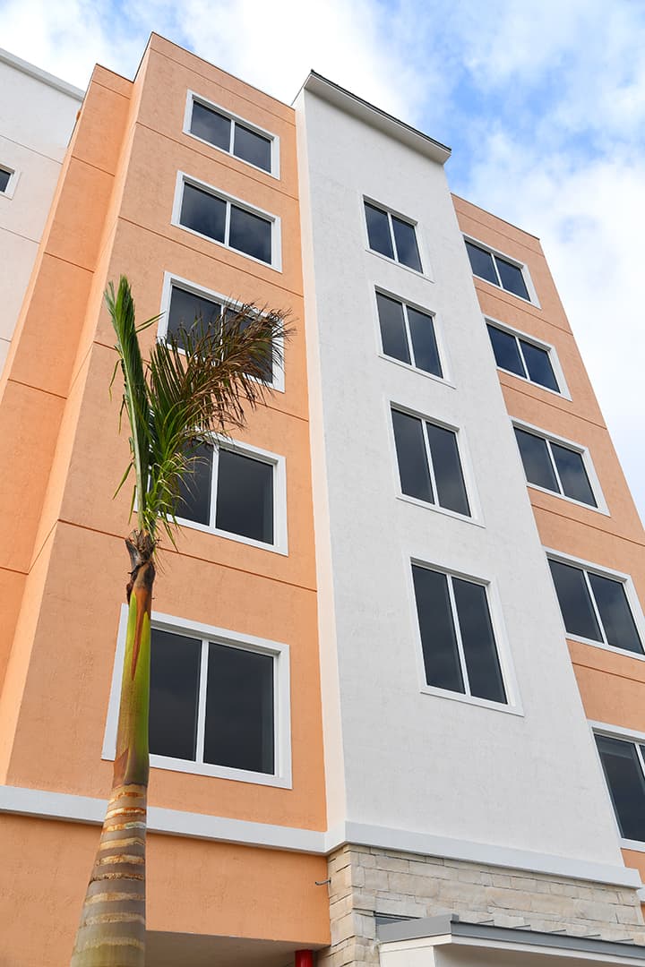 Orange and grey concrete building, six levels, two-panel impact glass with aluminum framing, FS-300 Impact Front Set windows by Aldora, palm tree in foreground