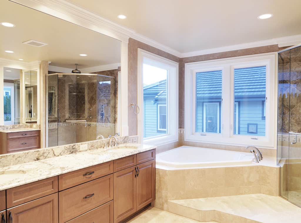 A bathtub is in the corner of the bathroom under windows. An Aldora mirror, sinks and a vanity cover the left wall. A walk-in glass shower is seen in the mirror reflection.
