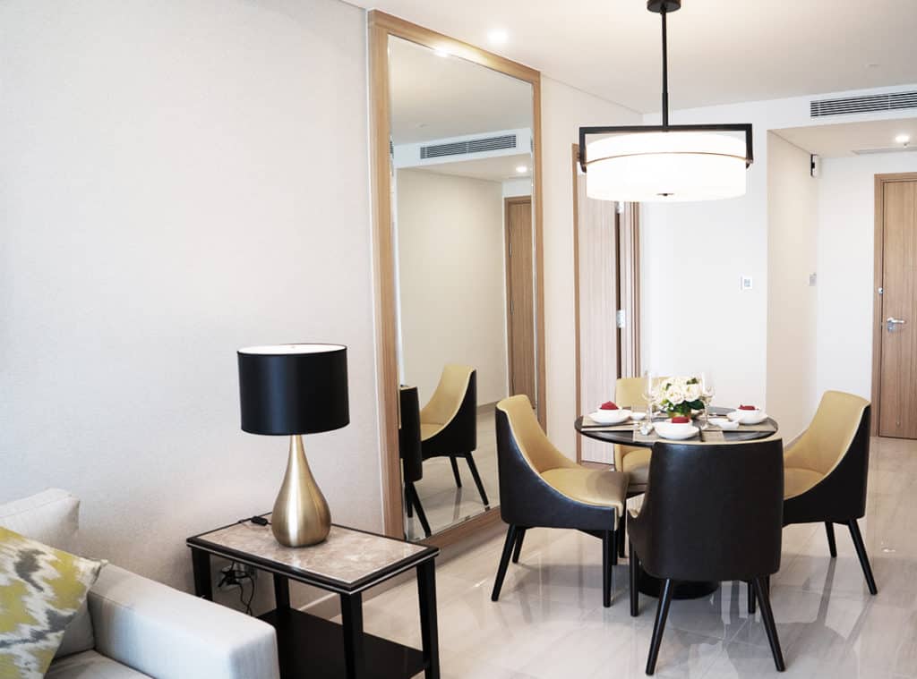 A set dining table with four chairs sits under a ceiling lamp, in front of a foyer and door. A floor to ceiling Aldora mirror covers the wall to the left of the table. A side table with a lamp sits next to a couch.