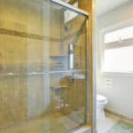 Aldora Fiesta glass shower doors see through to a tiled shower with shelves and hardware. A toilet, window and sink are seen to the right.