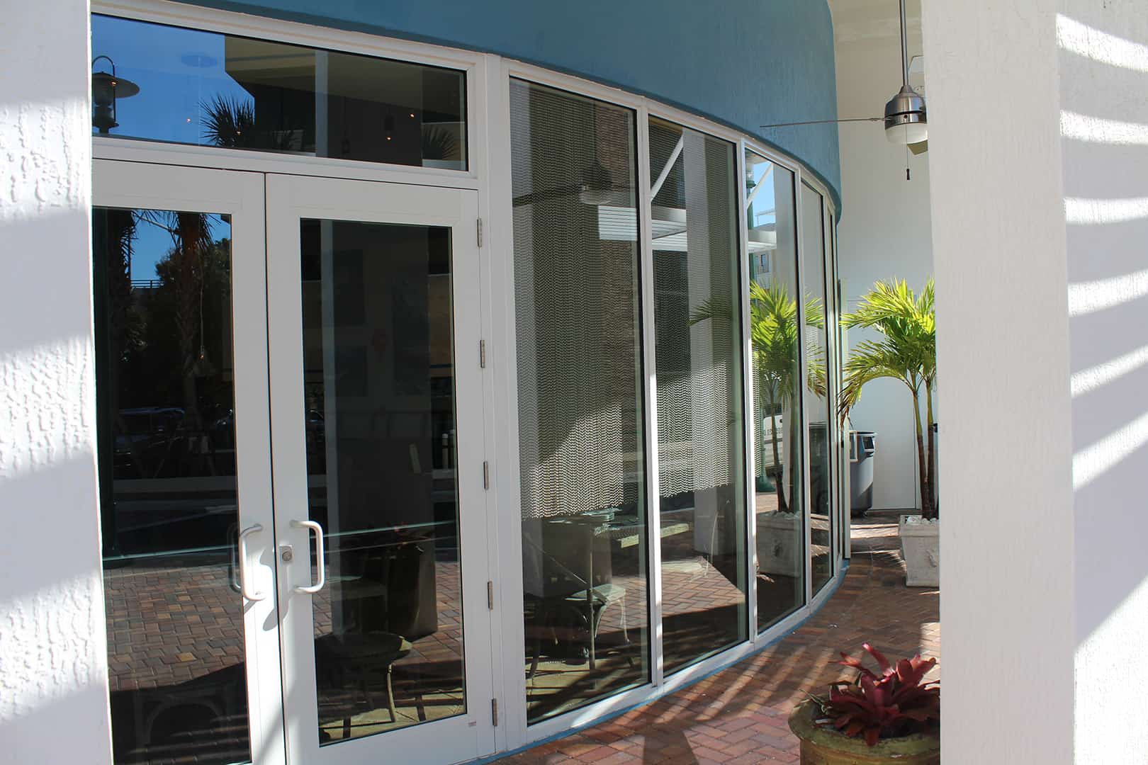 Curved outswing SMI-175 Impact Storefront System with Summit Impact Doors, floor-to-ceiling glass impact window, brick walkway, stucco exterior, tropical landscape to right of door and palm tree in background