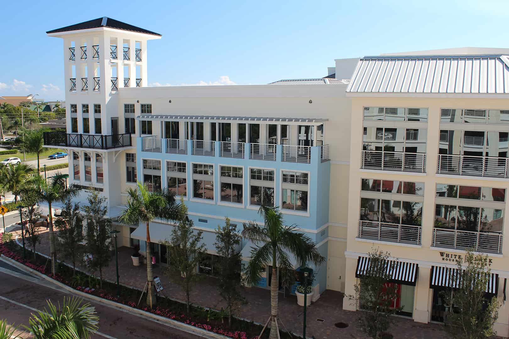 Multi-level beige storefront with blue accent wall, SMI-175 Impact Storefront System with Summit Impact Doors by Aldora, front-set impact windows and doors open to balconies on higher floors, tall white overlook on far left of building, palm trees line street