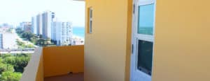 An exterior Aldora impact glass door is in view on the right wall of an orange building. A walled porch runs in front of the door and windows. Town landscape is seen in the background.