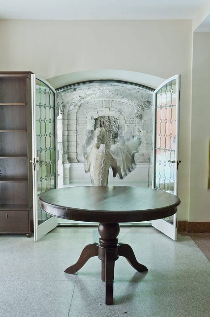 Internal entrance of arched glass double door with inlaid glass details, inward swinging glass door, round wood table in center of room, built in bookshelf on left