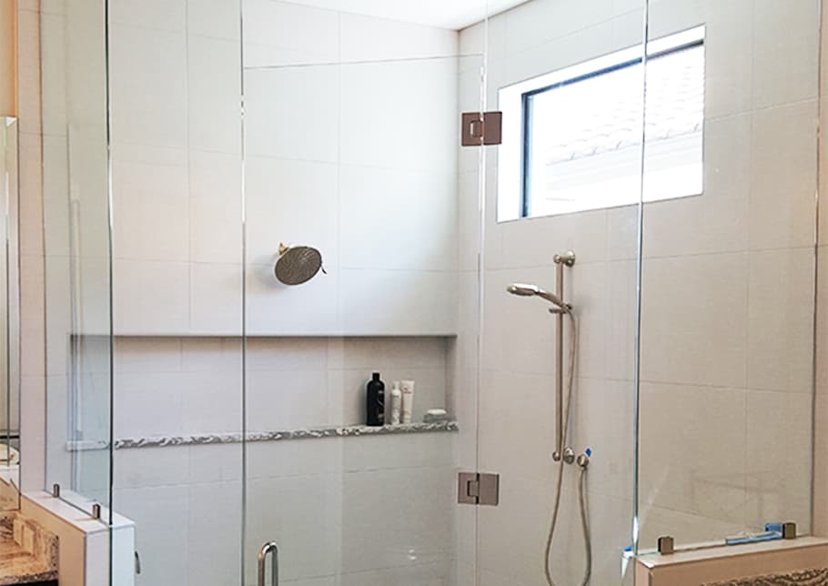 A shower enclosed in Aldora custom glass including the door. The tiled wall holds faucets, a shower head, a shelf and a window