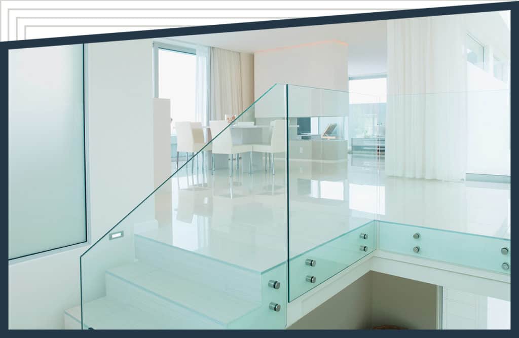 Aldora tempered glass acts as a railing on a staircase leading to a sitting room. White curtains line the windows, white chairs sit around a dining table and a white fireplace is beyond the table.