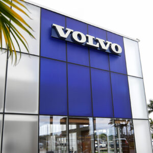 External view of Volvo car dealership, curtain wall glass with aluminum framing by Aldora, blue tinted glass on second level, front set system, manicured landscaping, parking lot and palm trees in background.