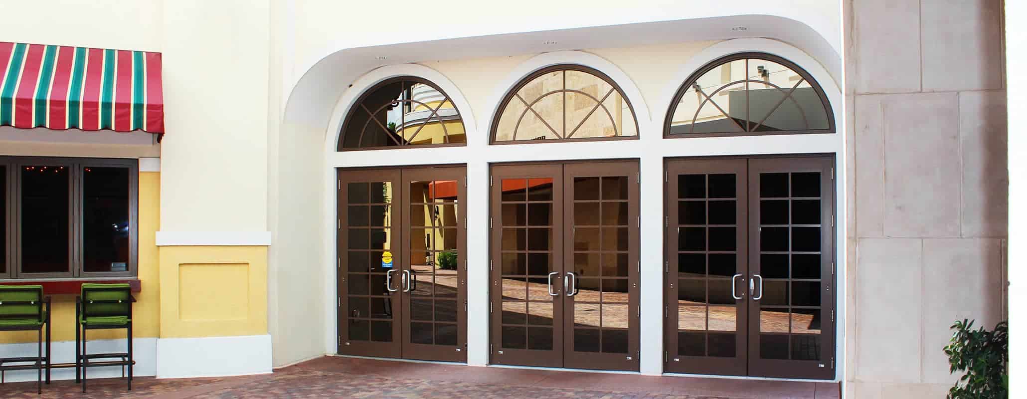 An exterior view of three Aldora impact glass doors with brick paved walkway in front and seating at the wall on the left.