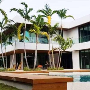 Two-story white home with glass impact walls, floor-to-ceiling insulated Aldora windows on first and second floors with black trim, SMI-175 Impact Storefront system from Aldora, grey accent pillars, infinity pool in foreground with wood trim, palm trees, tropical landscaping