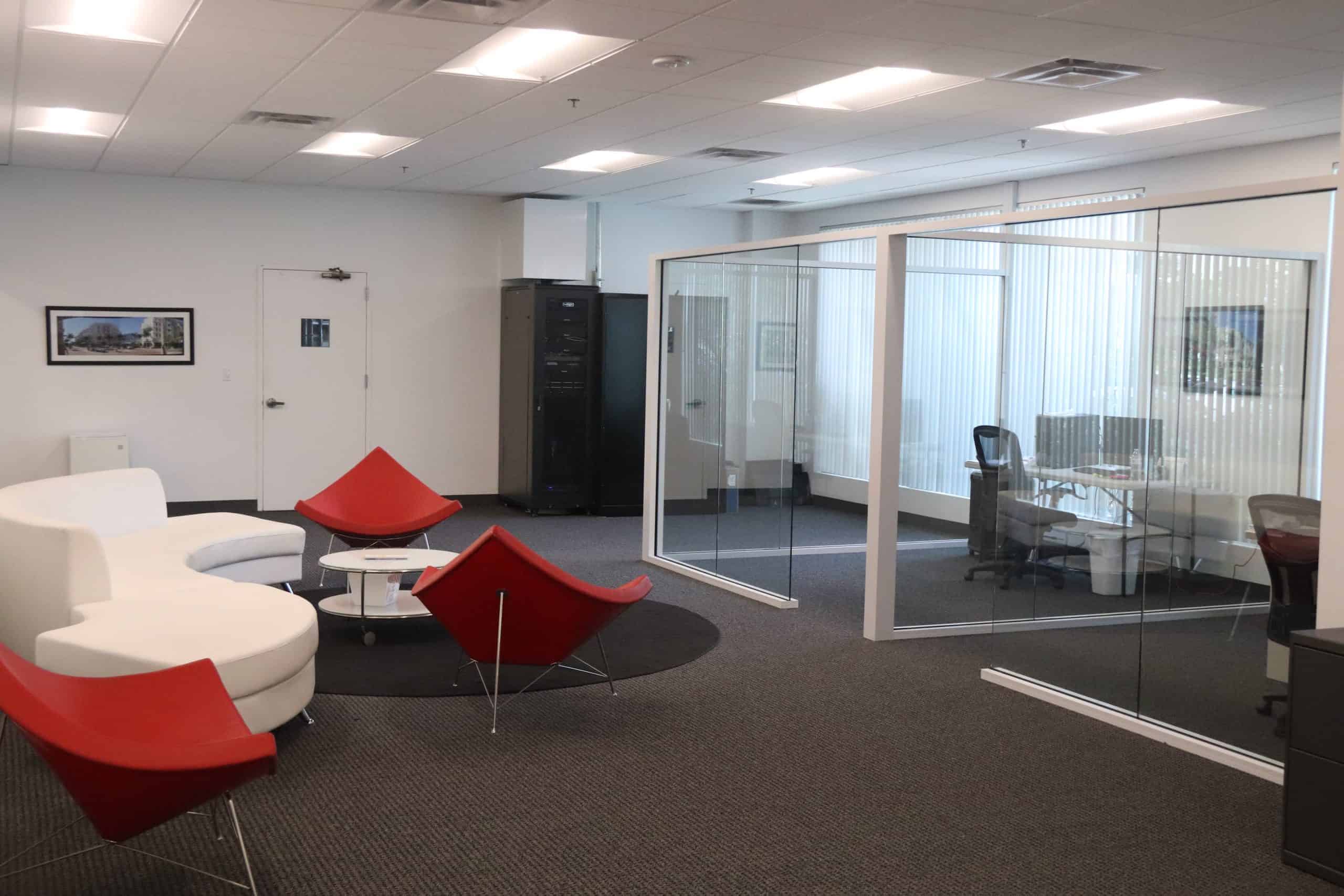 Side view of interior glass office wall partitions with office setups inside with white and red chairs in a lobby area.