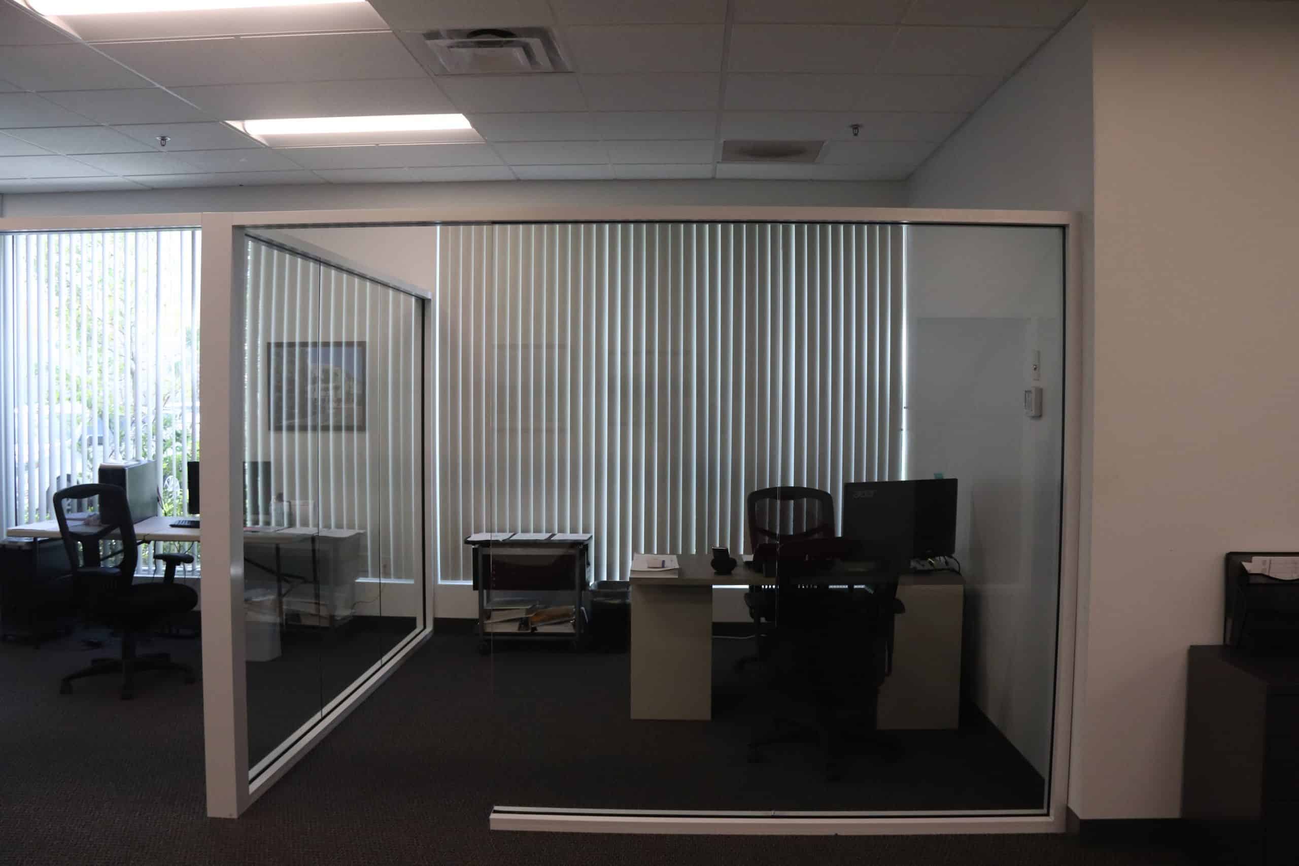 Interior glass office wall partition surrounds a desk with computer and chairs.